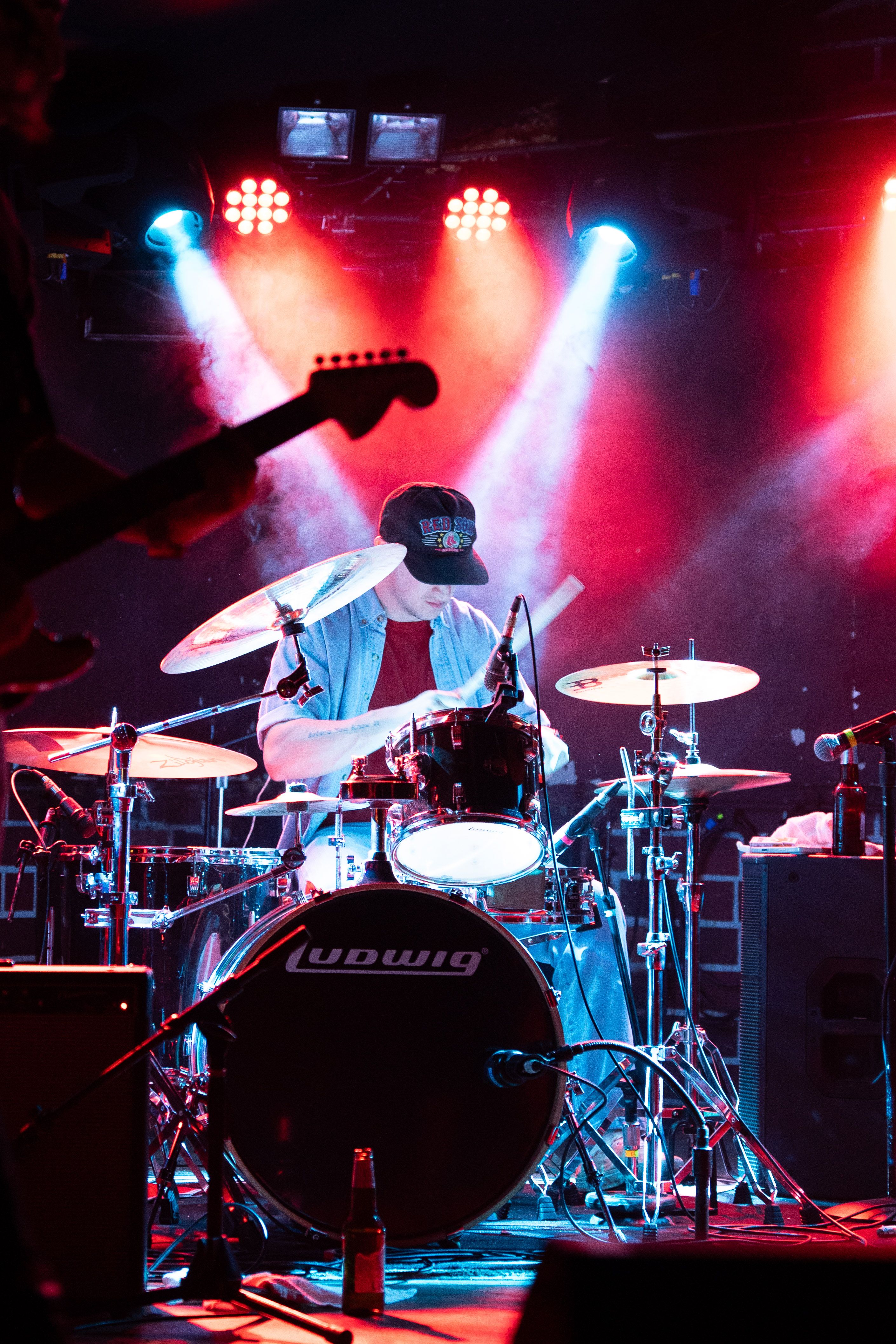 A drummer on stage, looking down and wearing a black hat. Two spotlights shine directly on him.